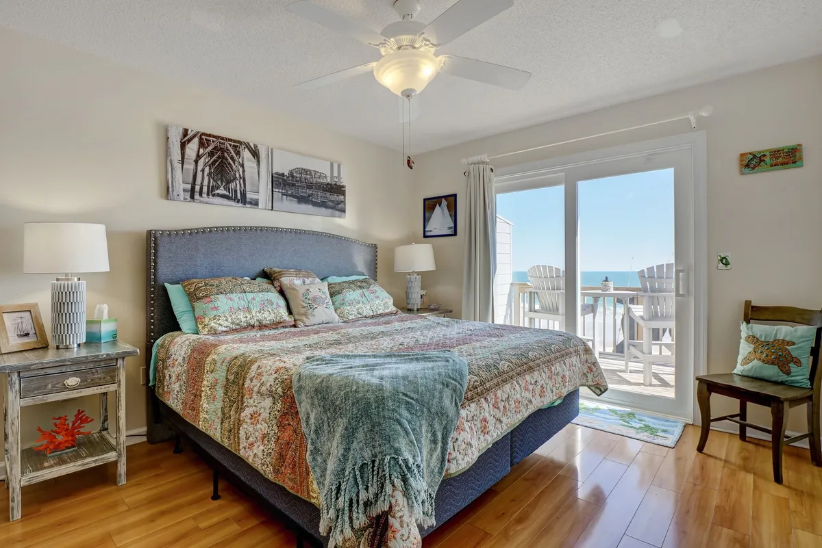 A serene upstairs bedroom with a large bed facing a sliding glass door that opens to a private balcony. The ocean view is prominent, and a nightstand with a lamp and tasteful coastal decor enhances the tranquil atmosphere.