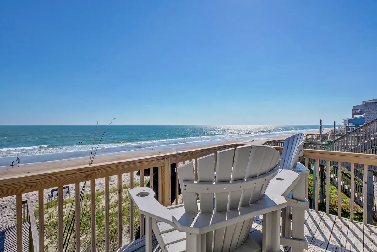 A pair of Adirondack chairs positioned on a deck overlook the beach, dunes, and ocean. The image suggests a peaceful retreat where guests can relax and take in unobstructed ocean views with their pets after a beach outing.