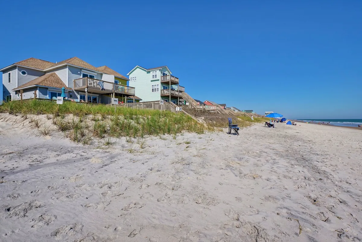 A bright exterior shot of beachfront houses lined up behind sand dunes, with beach chairs and an umbrella set up on the sand. The scene shows families gathered on the shore, with the ocean and expansive sky in the background. The setup suggests a day filled with beach activities, relaxation, and group fun right outside the house.