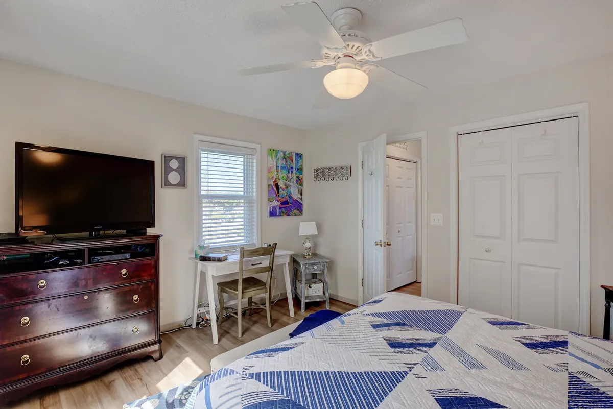 This image showcases a cozy bedroom with a patterned quilt, a television mounted on the wall, a dedicated desk with a chair, and colorful wall art. The window allows natural light to fill the room, and the workspace is clearly visible, making the area feel ideal for both rest and productivity.
