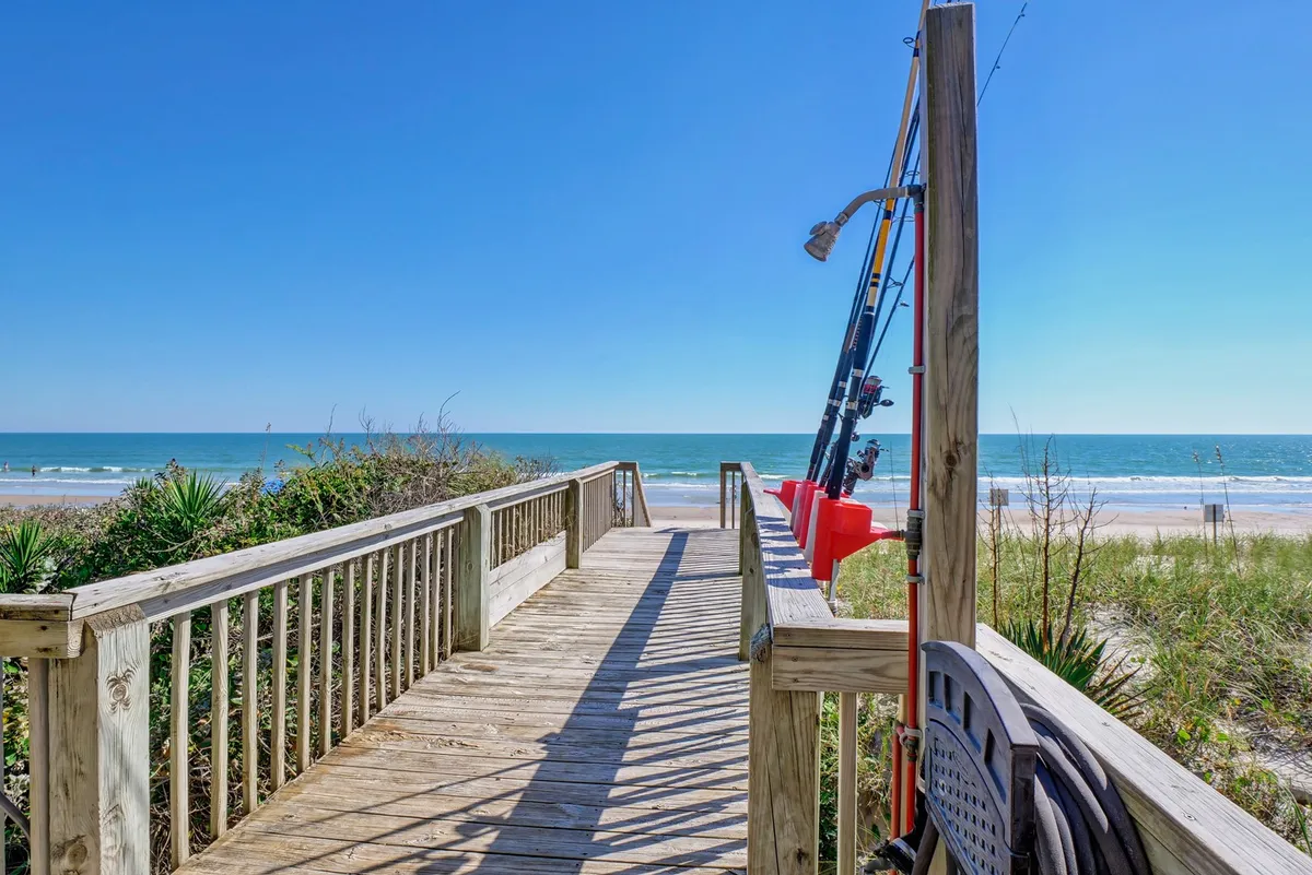 A wooden boardwalk stretching toward the ocean, with fishing rods visible and the beach in the background. The scene suggests convenient and direct access to the sand and surf, inviting exploration and play for both pets and owners.