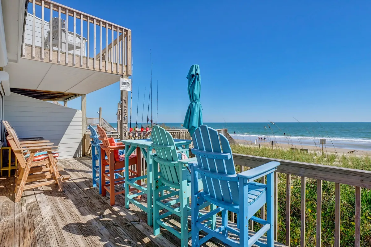 The hero image showcases a row of colorful Adirondack chairs on a deck facing the ocean, with a beach umbrella providing shade. The sandy beach and rolling waves are visible beyond the railing, inviting guests to relax, socialize, and enjoy breathtaking sunsets right from the comfort of the house.
