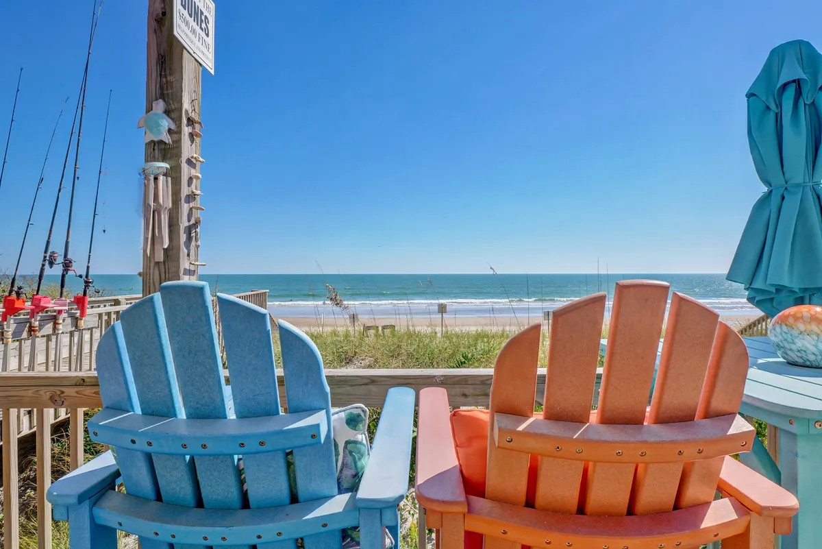 This image showcases a pair of colorful Adirondack chairs positioned with a sweeping ocean view in the background, fishing rods set up nearby, and a beach umbrella adding a splash of vibrancy. The overall mood is tranquil and cheerful, capturing both the inviting outdoor seating and direct proximity to the water.