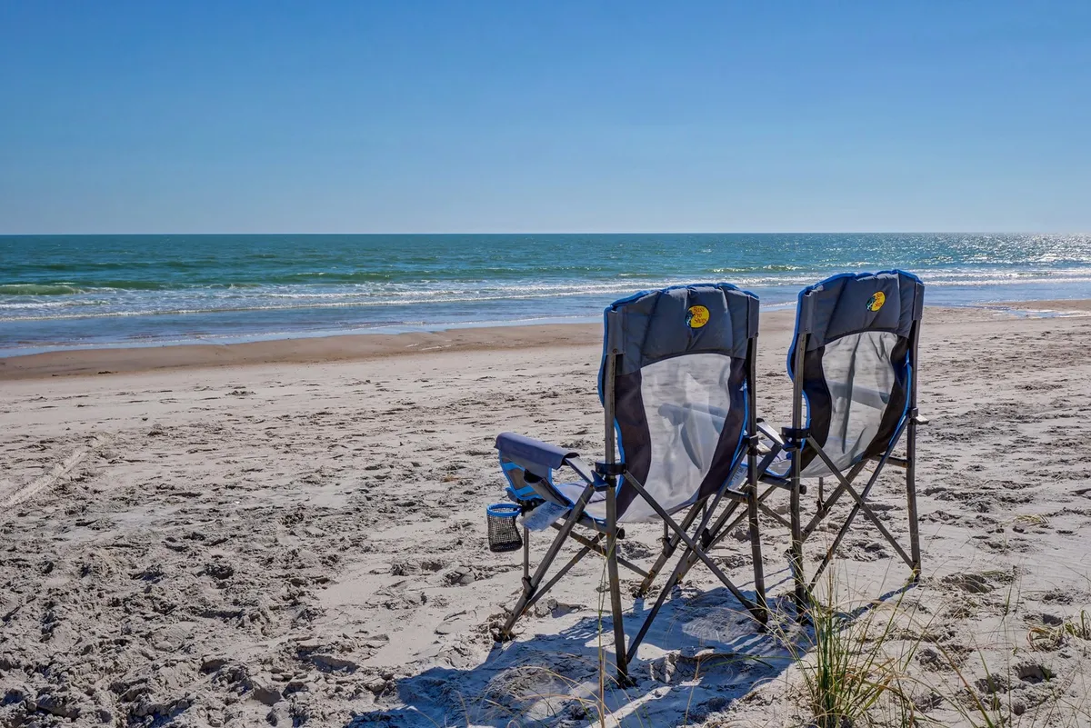 A serene exterior scene showing beach chairs set up on the sand, facing the ocean and wide blue sky. The image evokes the relaxation and leisurely group activities described in the section, such as sunbathing, socializing, and beachcombing together just steps from the house.