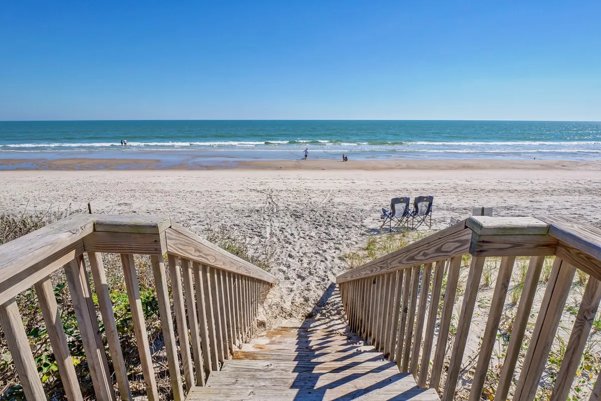 Wooden stairs descend from the beachfront deck directly onto soft sand, leading the eye toward empty beach chairs and rolling ocean waves. The image conveys a seamless transition from the comfort of the deck to the tranquility of the unspoiled shore below.