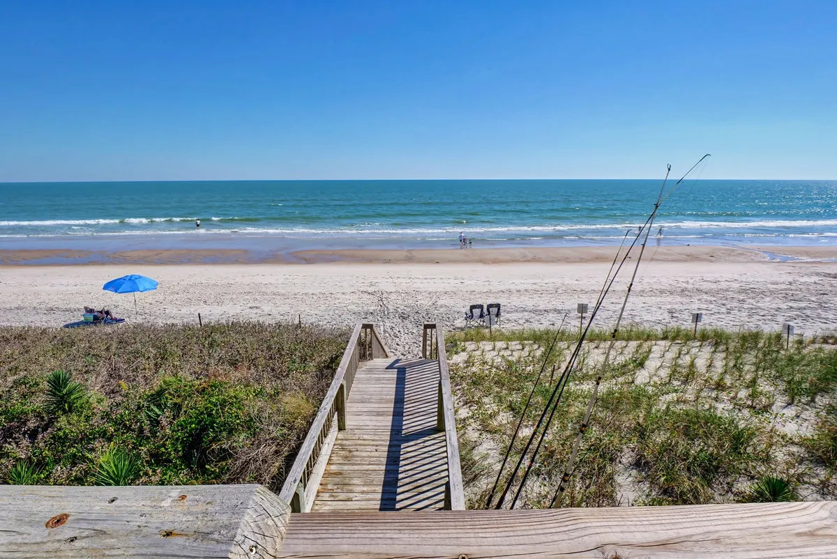 A wooden walkway leads directly onto the sandy beach, with a beach umbrella, gentle waves, and beachgoers visible. Sea grass frames the path, and the mood is peaceful, inviting viewers to imagine stepping outside and quickly reaching the shore.