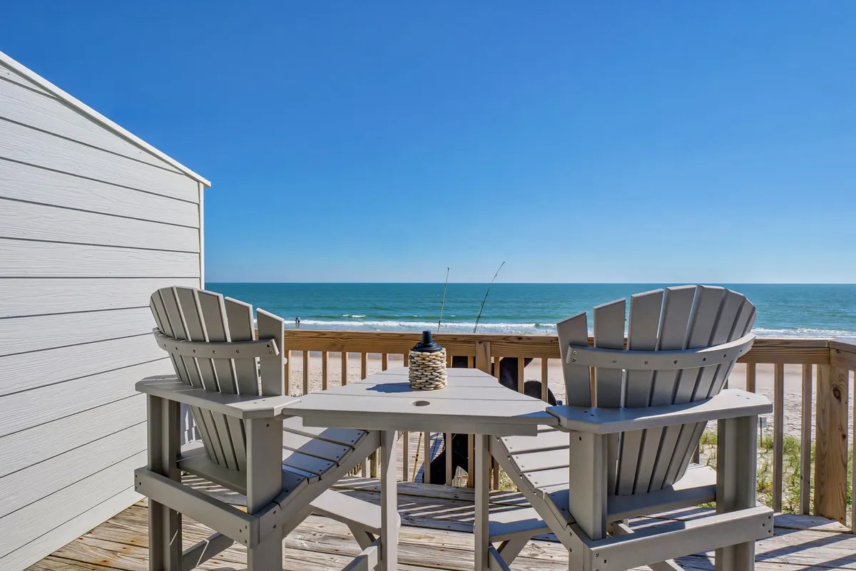 A tranquil exterior shot of a deck outfitted with Adirondack chairs and a small table, overlooking breathtaking beach views. The scene is set for unwinding with a drink, reading a book, or watching the sun set over the ocean.