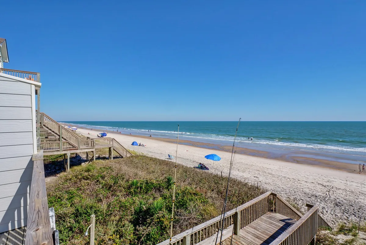 A tranquil view of the beach and ocean with a sunny sky overhead, a boardwalk leading toward the sand, and colorful umbrellas dotting the shore. Families and individuals are seen sunbathing, swimming, and strolling along the unspoiled beach.