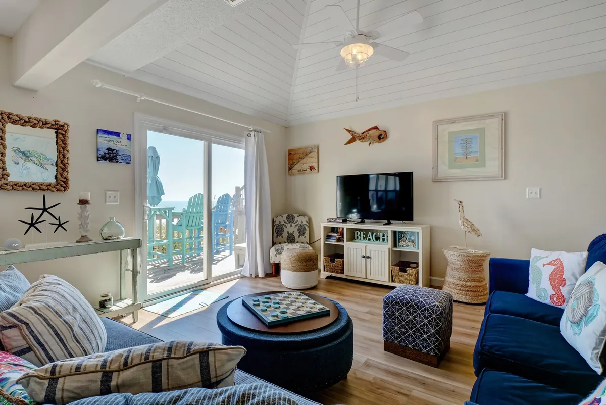 This image showcases a cozy living area with a comfortable sofa and coffee table facing a television. The room’s standout feature is the sliding glass door that opens directly onto the deck, providing a clear visual and physical connection to the outdoors. Sunlight streams in, hinting at the natural beauty waiting just outside.