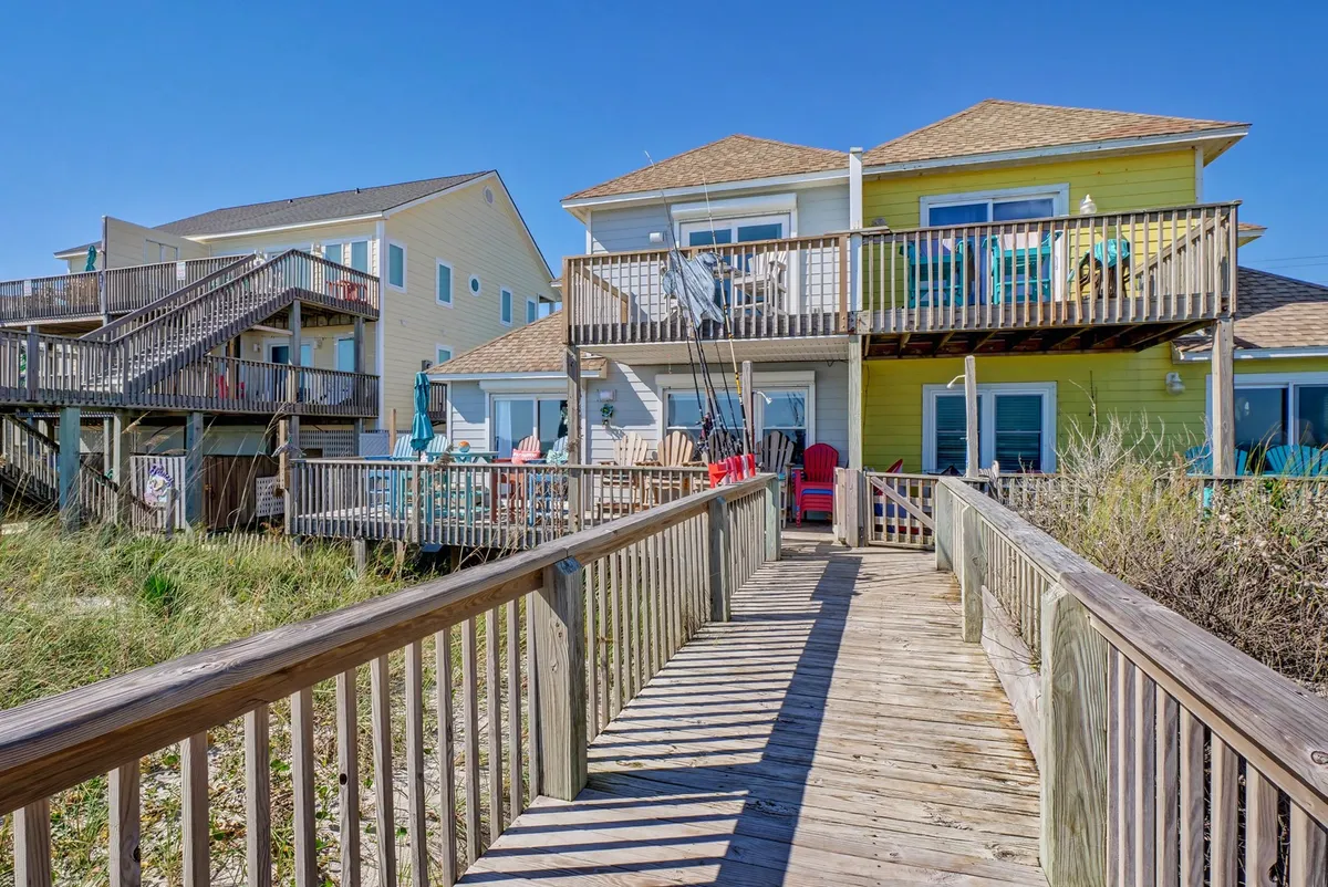 A lively beachfront exterior with a wooden walkway leading up to upper and lower decks. Colorful Adirondack chairs and balconies are visible, suggesting inviting spaces for relaxing, socializing, or enjoying the ocean views. Beach gear and cheerful details evoke a family- and pet-friendly atmosphere.