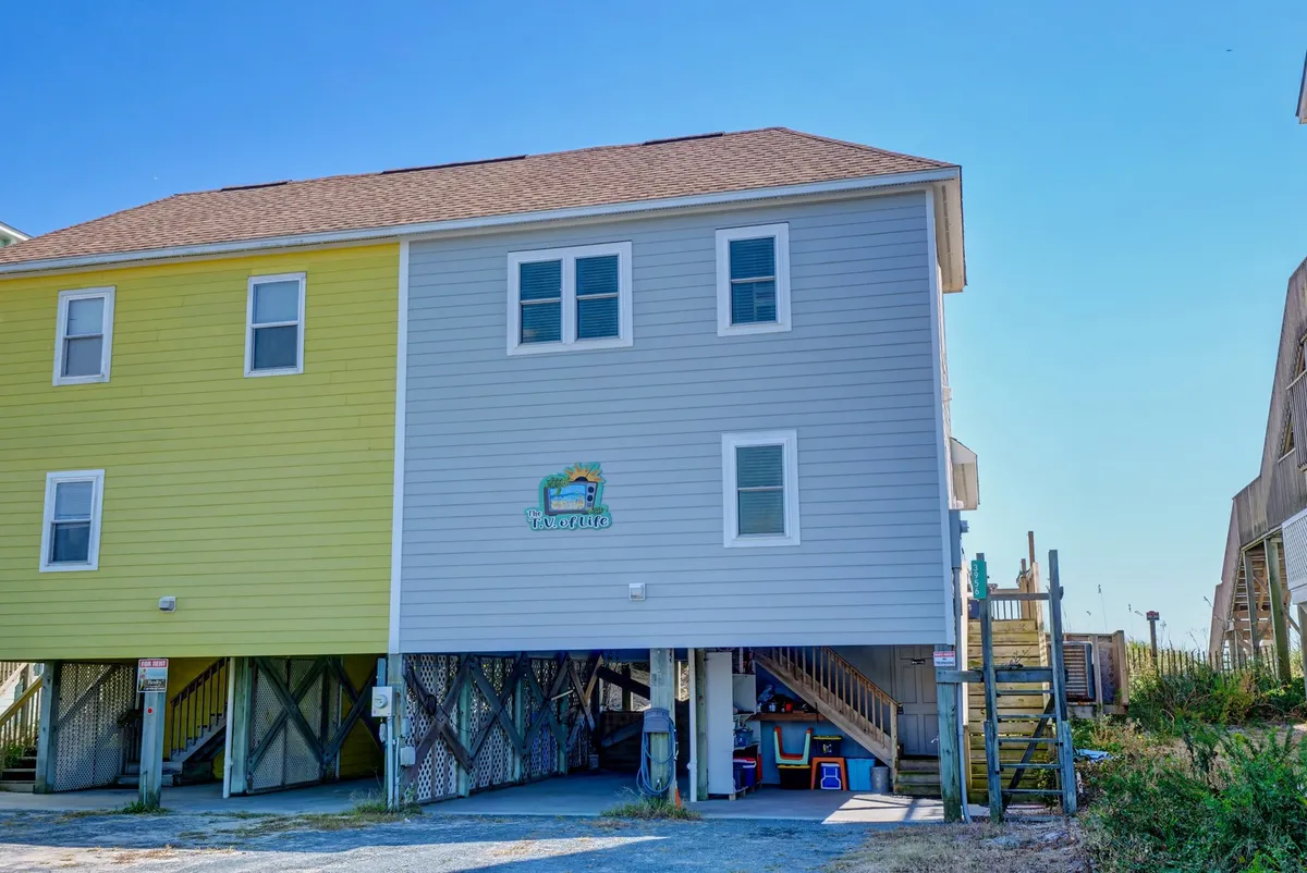 The exterior of a classic beach house is shown with two-tone paint, a visible 'Beach House' sign, and under-house parking. The image also hints at a deck space suitable for outdoor coffee or relaxation, set against the backdrop of a relaxed, coastal atmosphere.