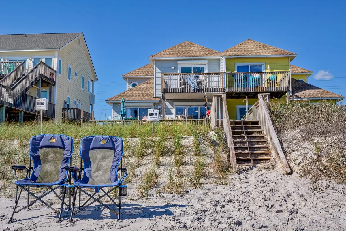 An inviting scene featuring beach chairs set up near a beach house, with a sand dune and stairs leading directly from the home to the shore. The setup suggests readiness for family gatherings, relaxing on the deck, and quick transitions from home to sand.
